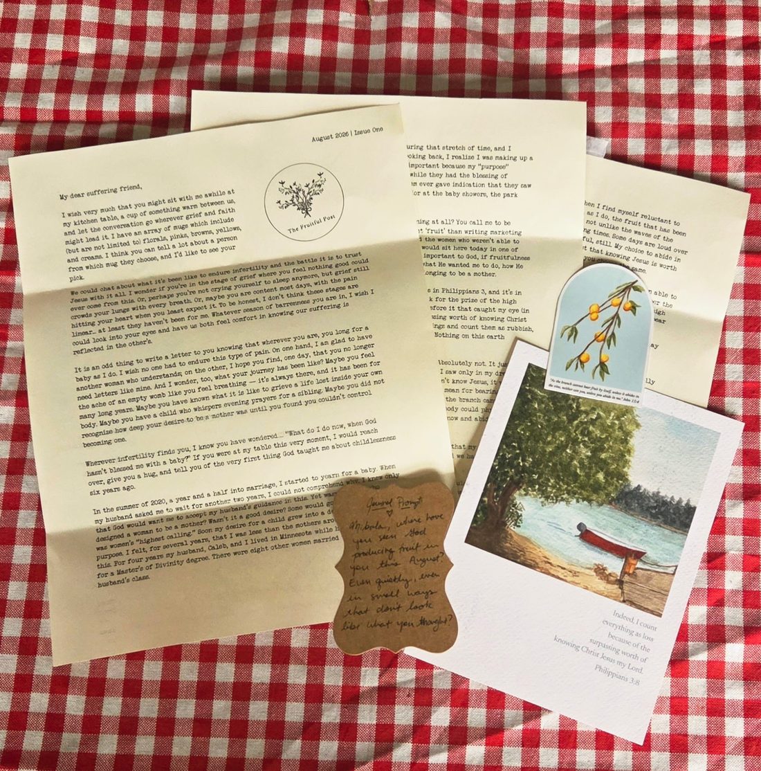 August's mailing laid out on a red gingham cloth: the typewritten letter, an art print of a red boat under a tree, a fruit-branch sticker, and a handwritten journal prompt card.