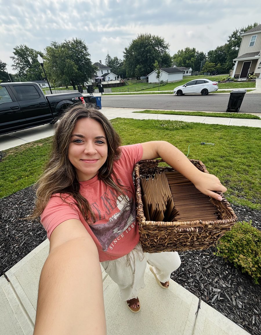 Jules standing in her driveway, holding a woven basket filled with kraft envelopes ready for the post office.